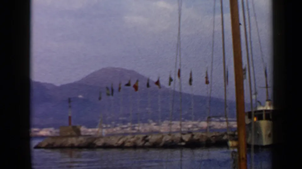 1960 8mm coastal harbor view showing a line of flags, sailboat mast and rigging, calm water, and mountains beyond the shore.