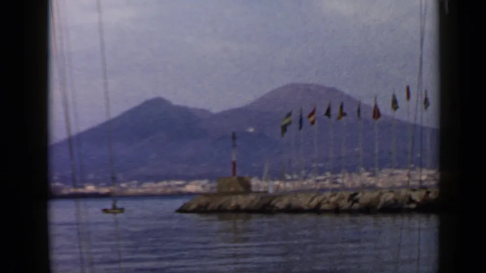 1960 8mm Italy harbor scene with calm water, a flagged breakwater, sailboat rigging, and hazy mountains framed by dark vertical edges.