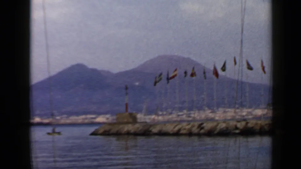 1960 8mm travel view of a bay in metadata-listed Rome, Italy, with flags, a rocky breakwater, small lighthouse, and hazy volcanic mountain.
