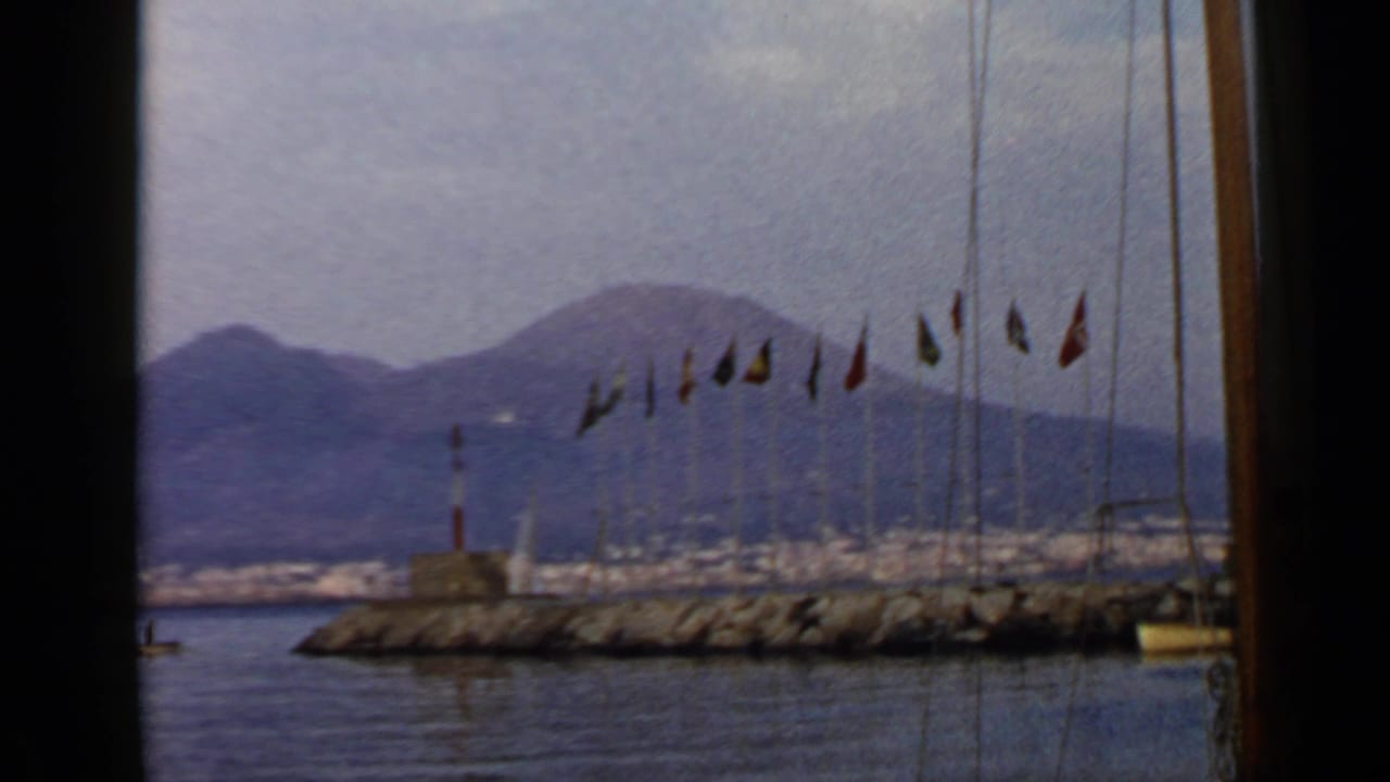 Metadata-listed Rome, Italy harbor scene on 8mm film, showing flags on a breakwater, a small lighthouse, calm bay water, and distant mountains.
