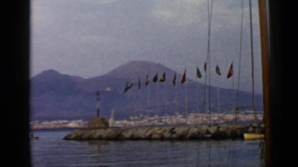 Metadata-listed Rome, Italy harbor scene on 8mm film, showing flags on a breakwater, a small lighthouse, calm bay water, and distant mountains.