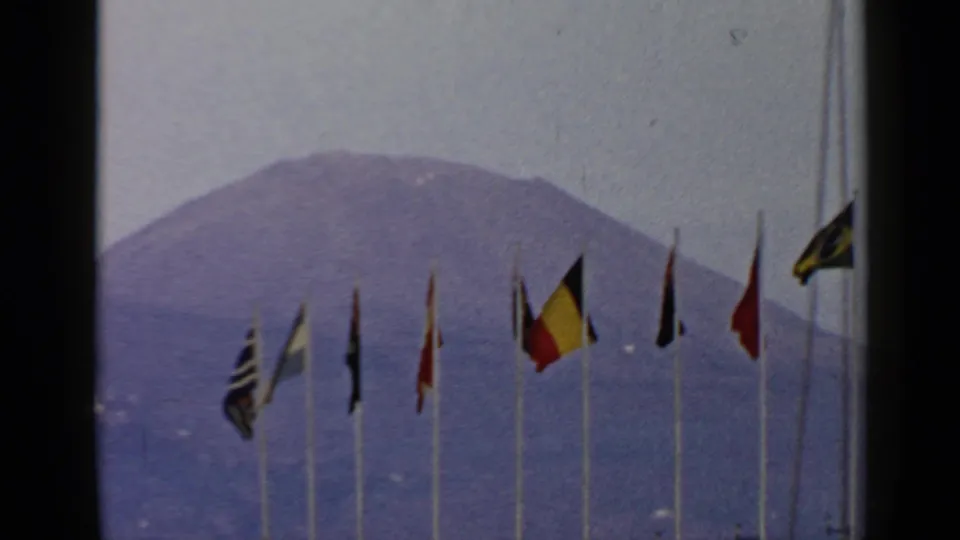 Closer 1960 8mm view of international flags in front of a hazy volcanic mountain, with the Italy location supplied by clip metadata.