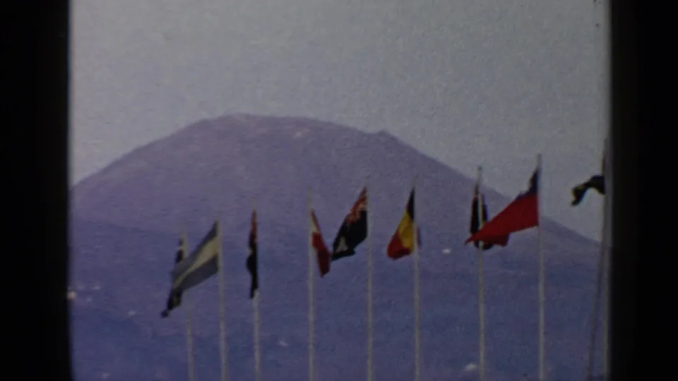 1960 8mm travel footage frame with national flags fluttering before a distant volcanic mountain near the metadata-listed Rome, Italy location.
