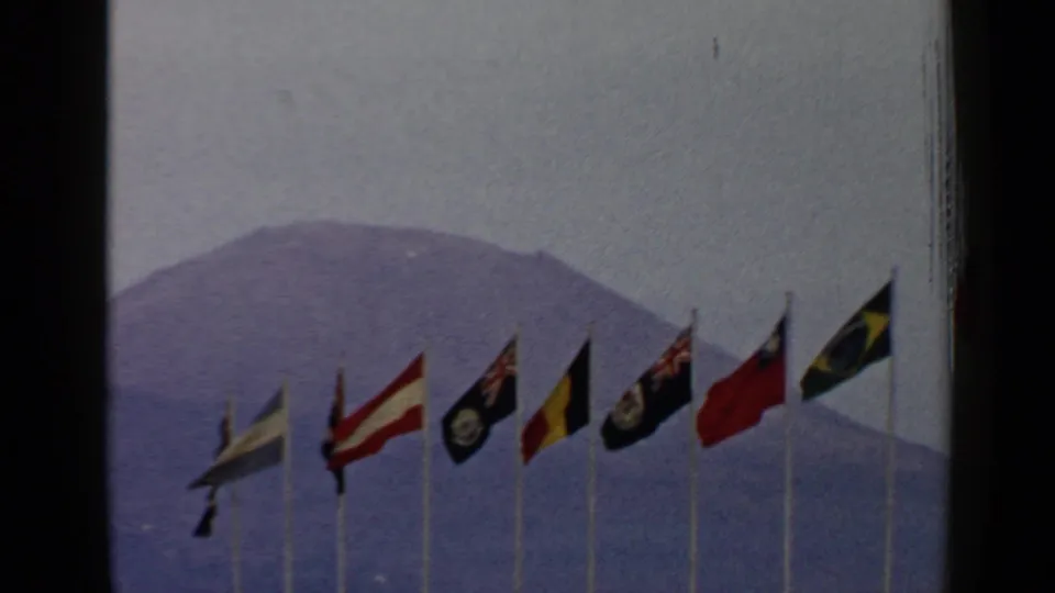 1960 Rome-area 8mm frame with several national flags on poles set against a hazy mountain.