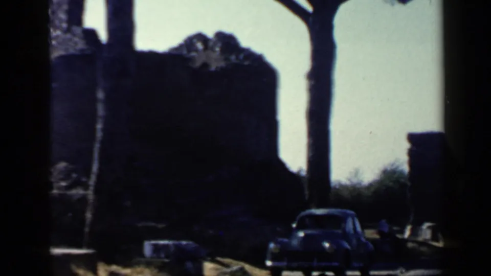 1960 Rome 8mm frame showing a dark stone ruin, tree trunks, roadside, and a parked period car.