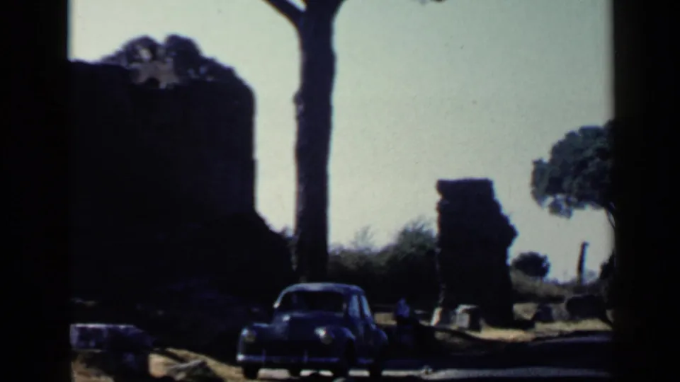1960 Rome 8mm frame with ruins, roadside trees, open sky, and a dark compact car parked nearby.