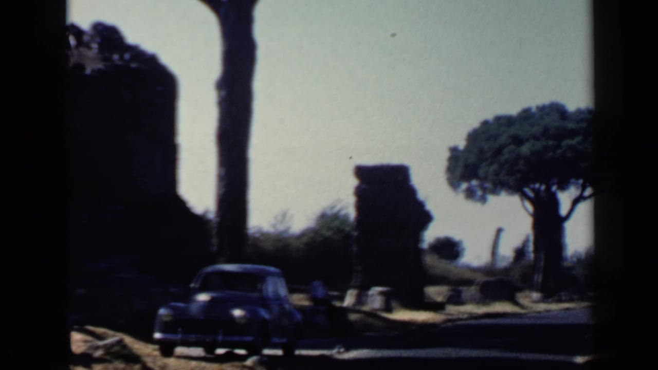 1960 Rome 8mm frame with a dark car at left, weathered ruins, bright sky, and a broad umbrella pine beyond the road.
