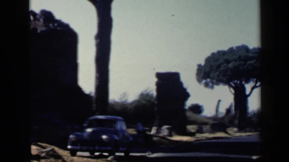 1960 Rome 8mm frame with a dark car at left, weathered ruins, bright sky, and a broad umbrella pine beyond the road.