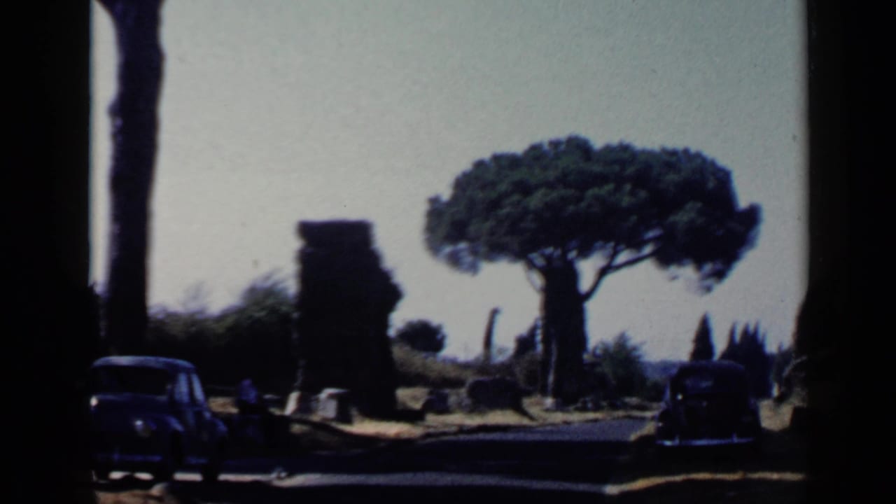 1960 Rome travel frame showing a dark car on a road between stone ruins, with an umbrella pine and bright open sky behind it.