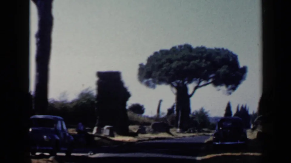 1960 Rome travel frame showing a dark car on a road between stone ruins, with an umbrella pine and bright open sky behind it.