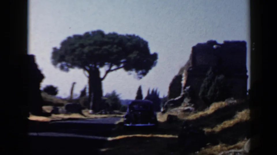 1960 Rome frame with the car low at right, a large umbrella pine centered, and weathered stone ruins framing the road.