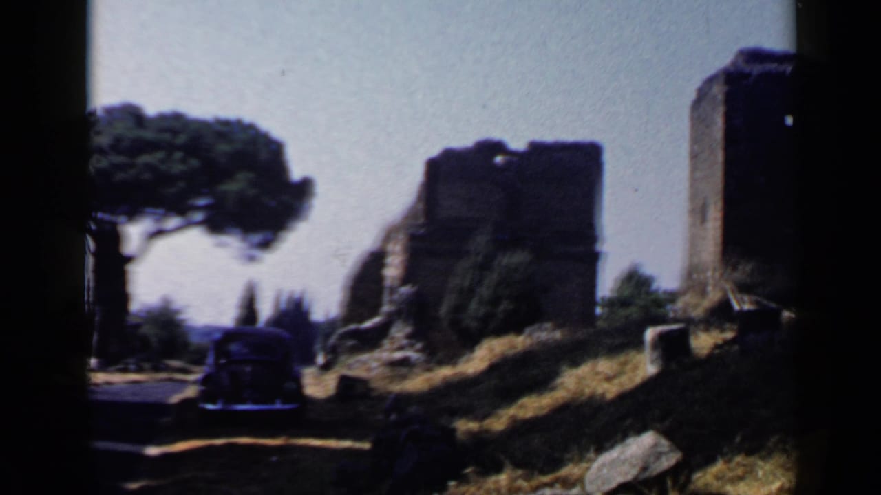 Roadside ruins with a large tree, dark car, dry grass, and rocks.