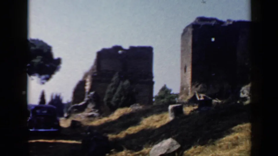 1960 Rome frame with a dark car at the lower left, stone ruins to the right, dry grass, rocks, and a tree partly cropped by the film edge.