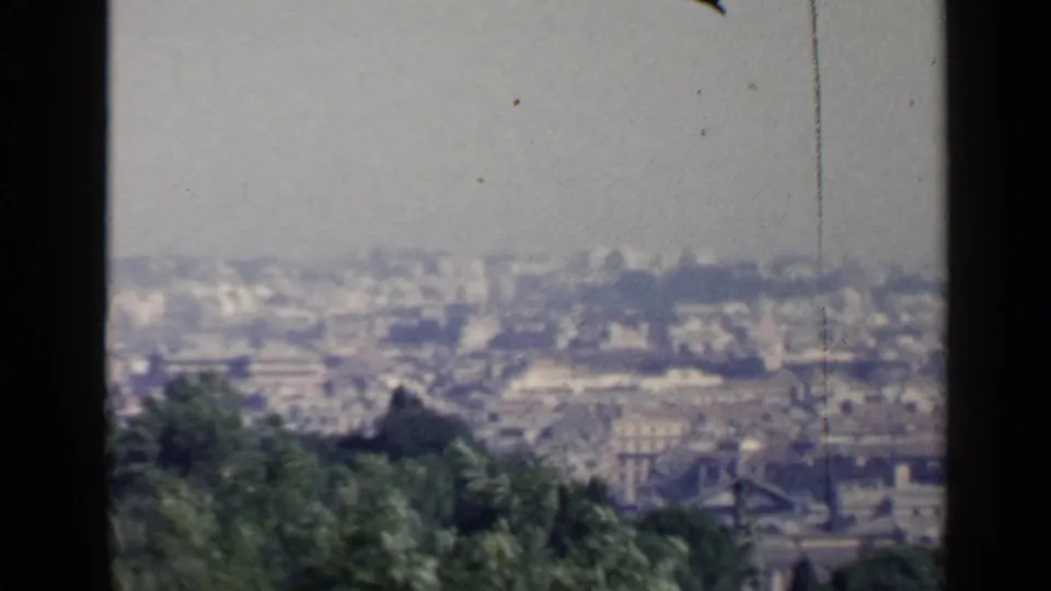 1960 8mm view from above Rome, with soft rooftops, haze, foreground greenery, and dark film-frame borders.
