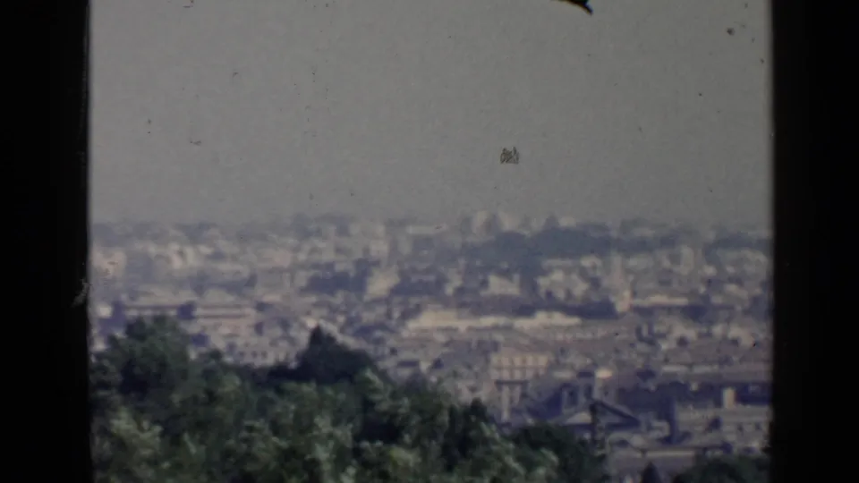 Soft 1960 home-movie frame looking across Rome from an overlook, with greenery below and a pale, hazy urban horizon.