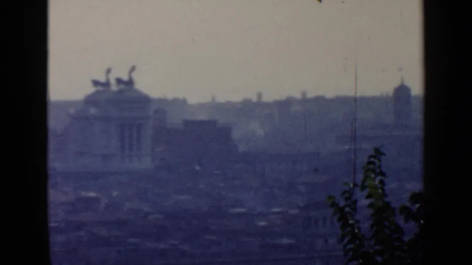 Rome skyline detail from 1960 8mm footage, showing a distant monumental building with rooftop statuary through blue haze.