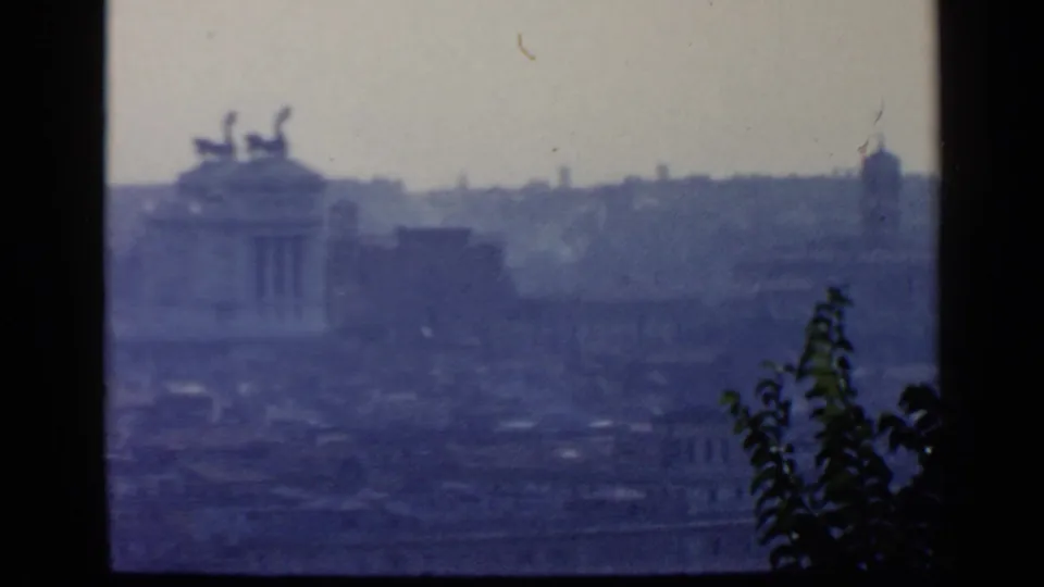 Elevated Rome view on 8mm film, with a distant monumental structure, dense rooftops, blue haze, and leafy foreground detail.