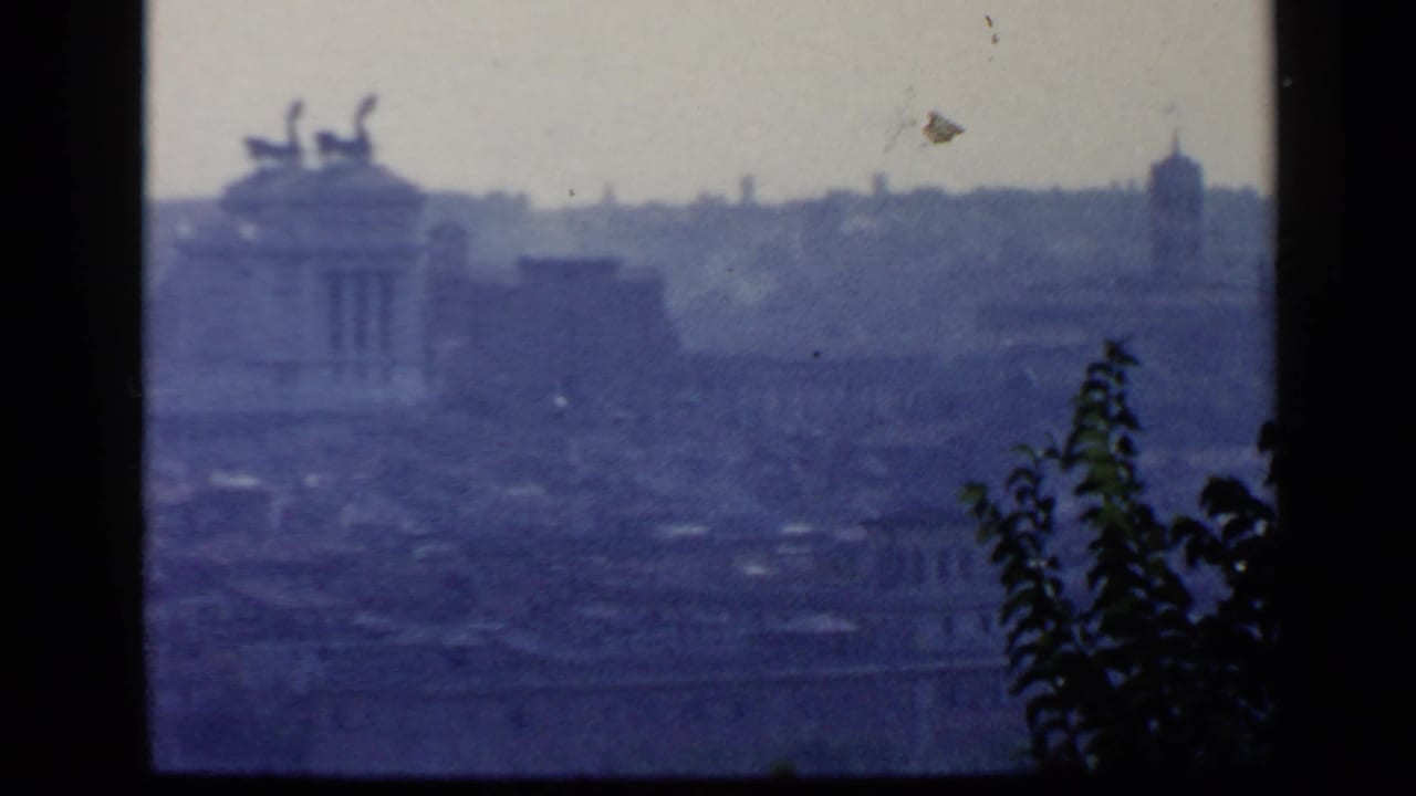 Hazy elevated view over Rome rooftops with a classical roofline at left and foliage near the center.