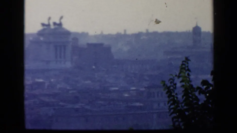 1960 8mm view over Rome from a high vantage point, with distant hills, dark frame borders, foliage, and a classical building roofline.