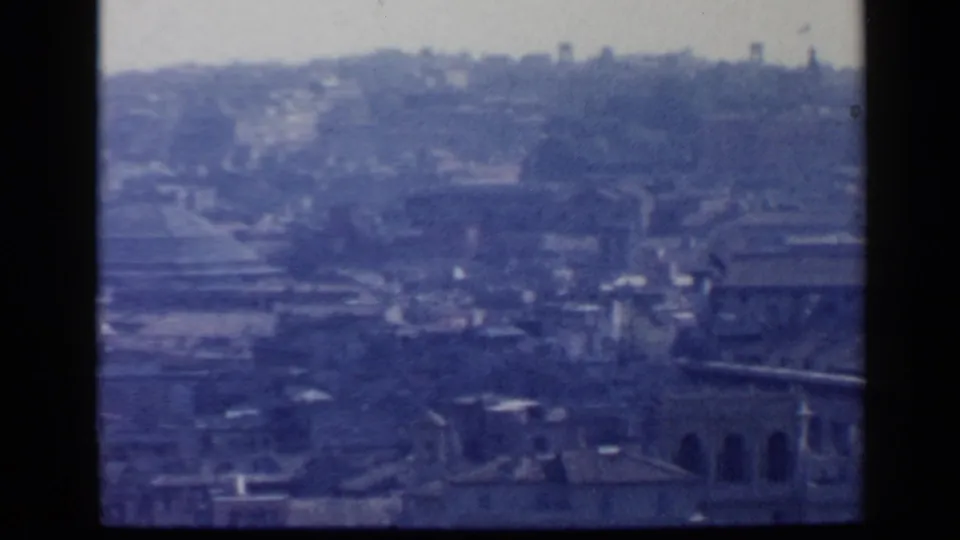 Rome appears from an elevated 1960 home-movie viewpoint, with layered rooftops, a distant hillside, and a hazy blue film cast.