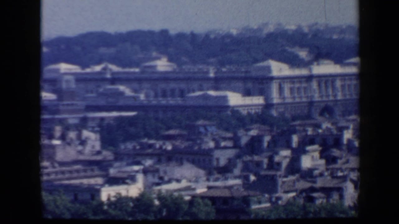 Elevated view over Rome rooftops with a long historic building and wooded hill beyond.