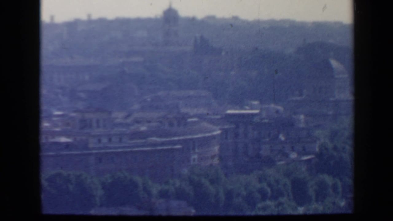 Hazy city view with stone buildings and a distant dome beyond trees.