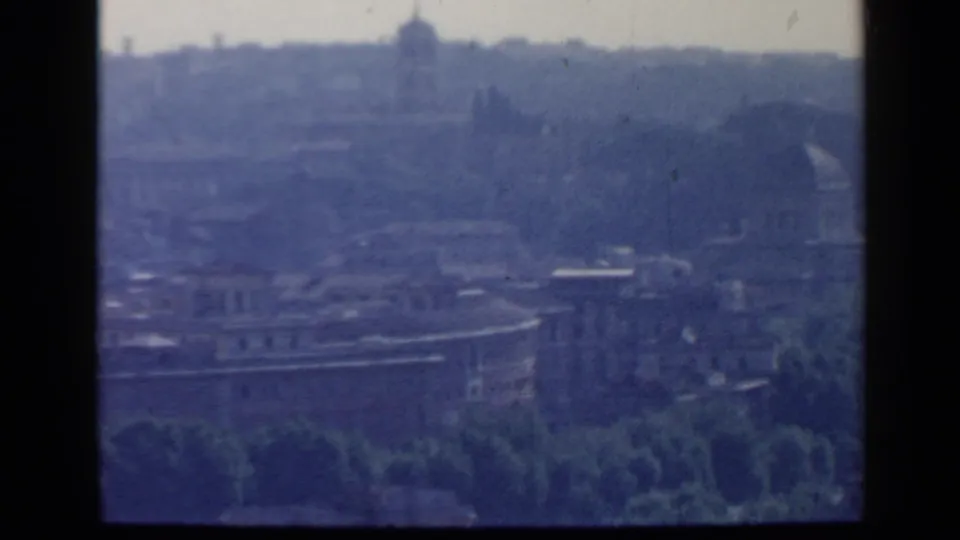 1960 Rome home-movie frame showing a soft, blue-tinted view of stone buildings, trees, and a distant dome.