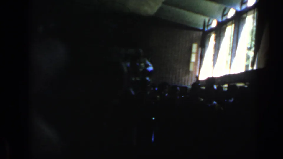 1960 Rome home-movie frame inside a shadowed hall, with bright windows and indistinct seated or gathered forms.
