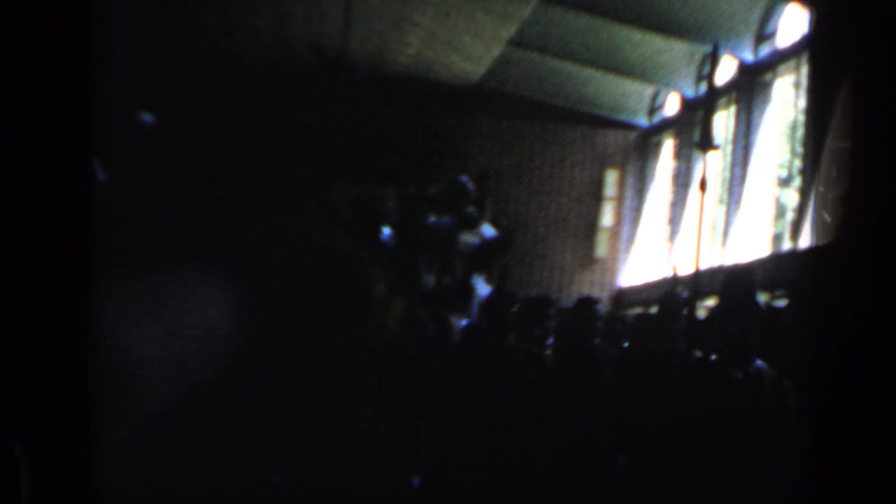 Dim hall with bright windows and silhouetted spectators around a carried seated figure.