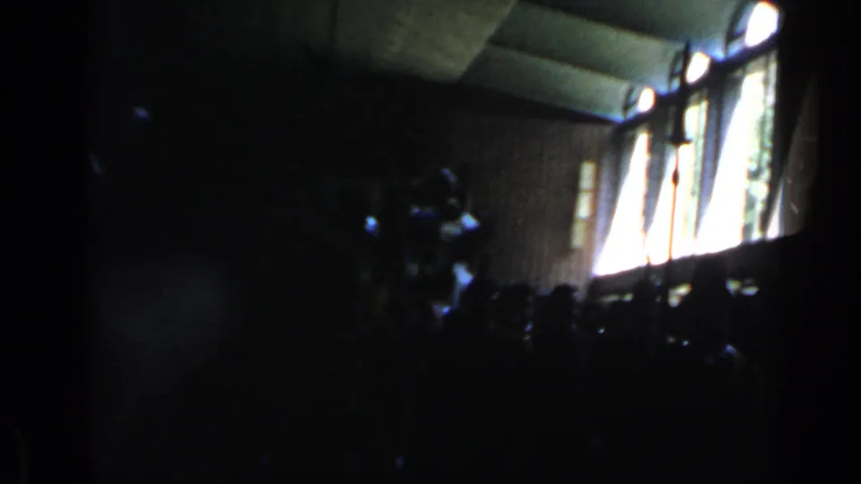 1960 Rome home-movie frame showing a dark interior hall, tall windows, and silhouetted people around a likely papal chair procession.