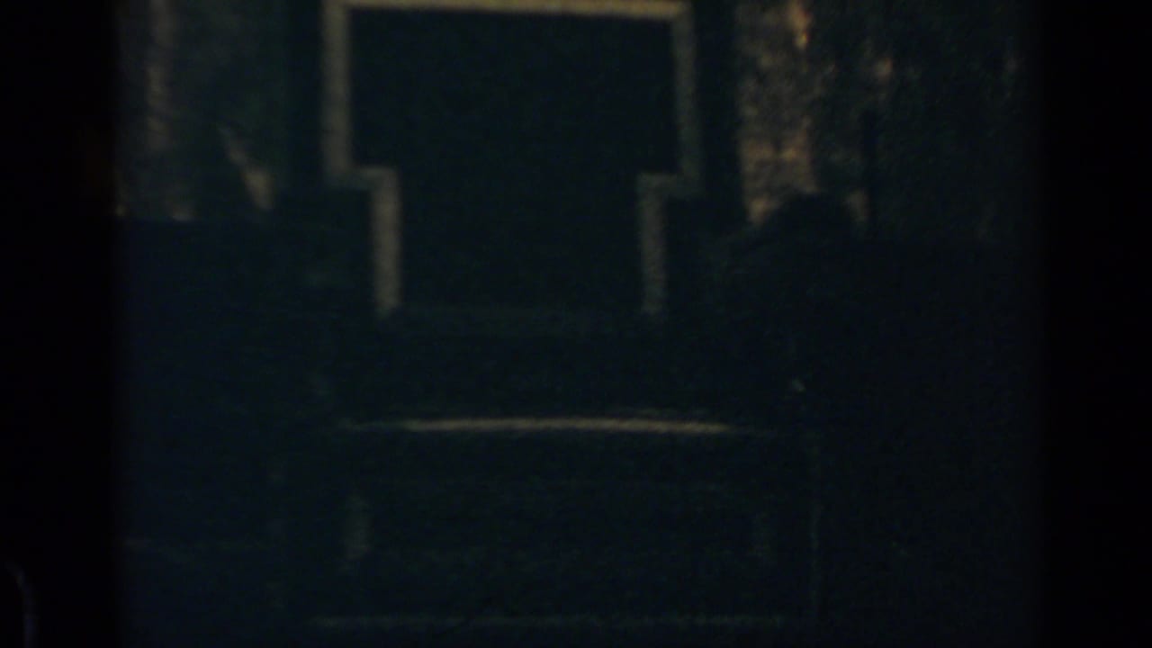 Dim 8mm frame from the Rome clip showing a shadowed facade and faint crowd forms near the lower edge.