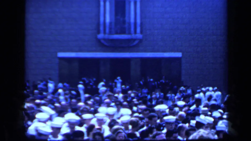 1960 Rome home-movie frame with a packed crowd outside a pale building, including many white hats and a tall arched window.
