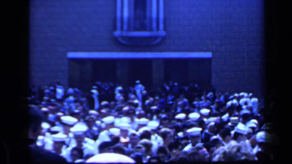 1960 Rome home-movie frame showing a dense crowd outside a stone building, with many people in white sailor-style caps near the entrance.