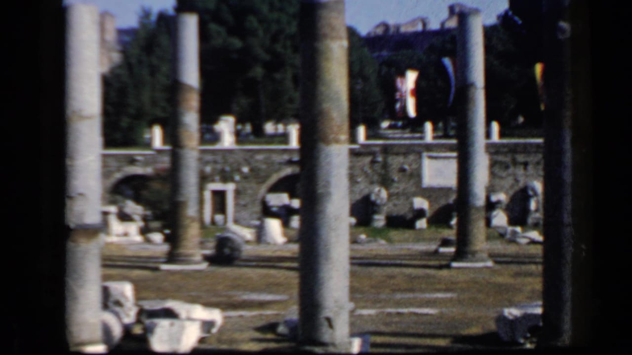 1960 8mm view in Rome showing ancient stone ruins framed by tall weathered columns, with arches, fragments, trees, and bright sky.