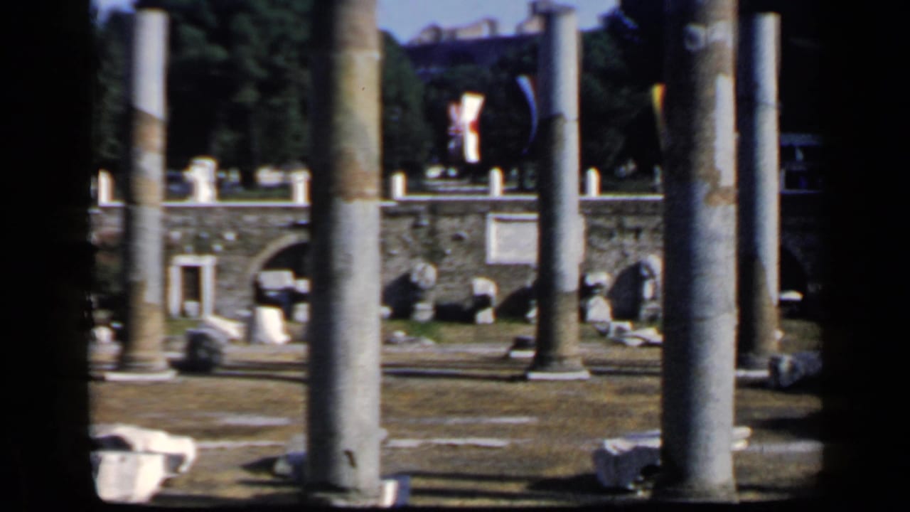 Rome ruin site on 8mm film, with tall foreground columns, arched masonry, scattered stone pieces, trees, and dark frame edges.