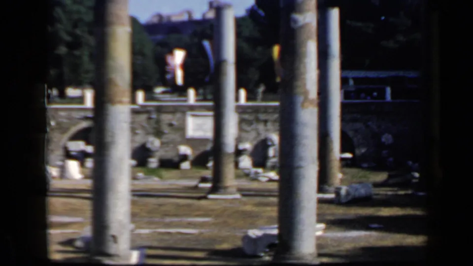 Vintage 8mm Rome footage shows weathered columns before ancient arches and scattered fragments, with soft focus and heavy edge shadowing.