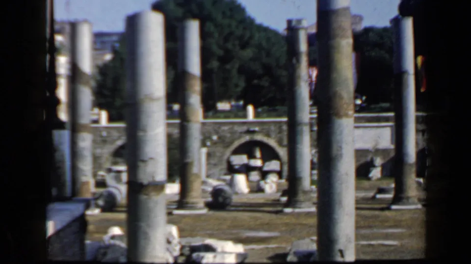 Final 8mm frame of Roman ruins, showing several foreground columns, a low arched wall, stone fragments, trees, and bright outdoor light.
