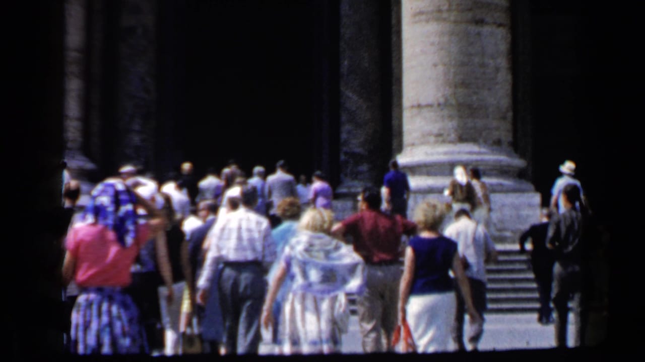 1960 Vatican home-movie frame with tourists approaching stone steps beneath massive columns and dark entryways.