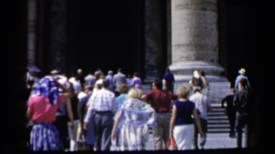 1960 Vatican home-movie frame with tourists approaching stone steps beneath massive columns and dark entryways.