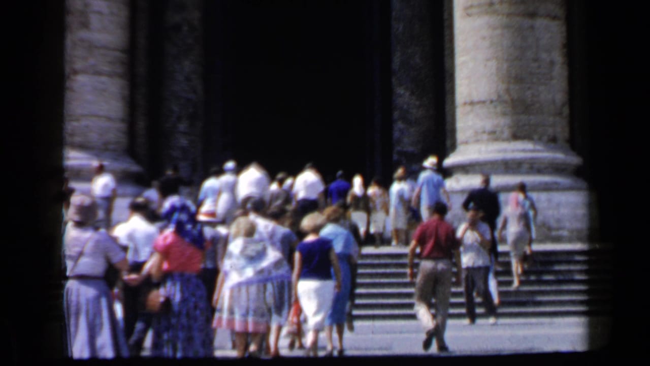 Tourists in summer clothing ascend Vatican stone steps framed by columns, with faces visible at a distance.