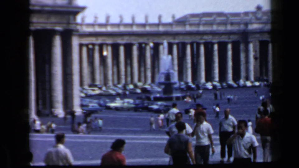 1960 8mm footage frame showing the Vatican colonnades, a fountain, parked cars, and visitors walking through St. Peter's Square.