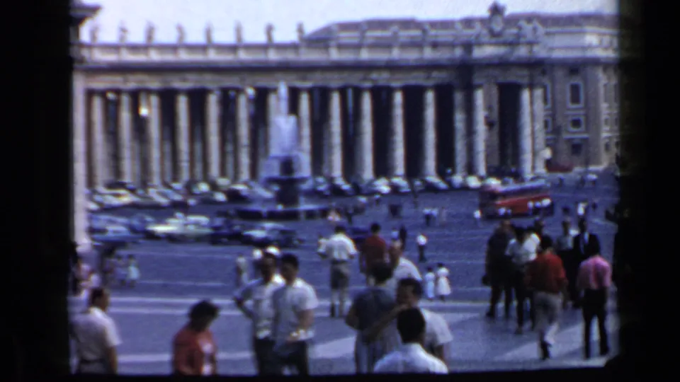 1960 Vatican City home-movie frame with St. Peter's Square colonnades, a fountain, parked cars, and visitors moving across the plaza.