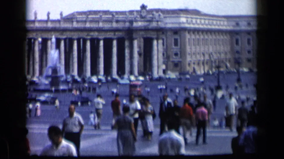 Visitors in Vatican City's St. Peter's Square walk past the camera while the colonnade, fountain, and cars appear in the background.