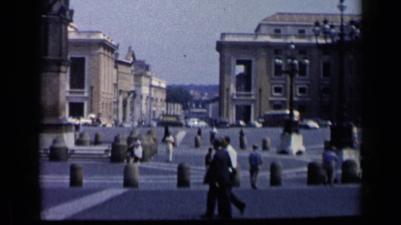 Public plaza with pedestrians, stone bollards, steps, cars, and classical buildings.