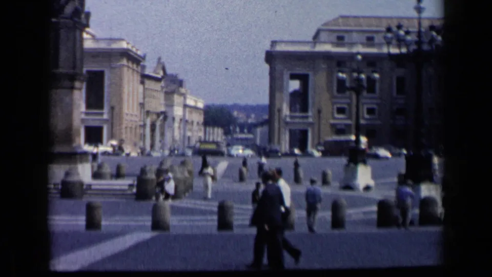 1960 8mm view of a Vatican City public plaza with pedestrians walking near stone bollards, steps, parked cars, and monumental buildings.