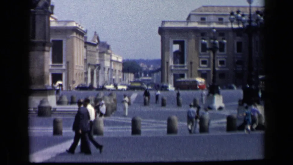 1960 8mm frame of pedestrians in a Vatican City plaza, with stone bollards, steps, parked cars, and large public buildings in the background.