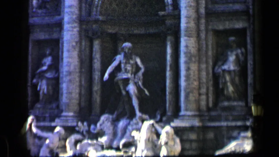 1960 8mm view of a monumental fountain with sculpted figures, stone niches, columns, water highlights, and visible film-frame edges.