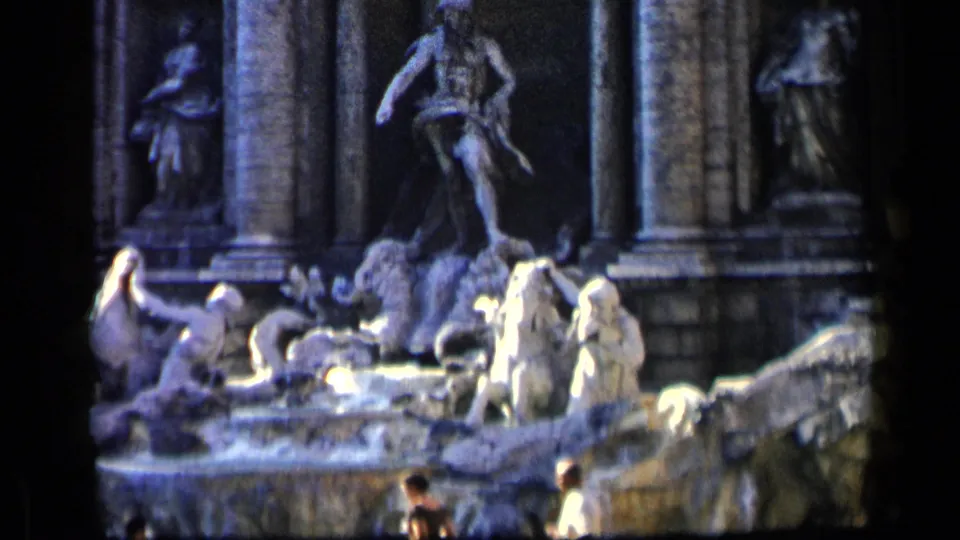 1960 8mm frame of the Trevi Fountain with bright stone figures, water, and two small visitors moving across the foreground.
