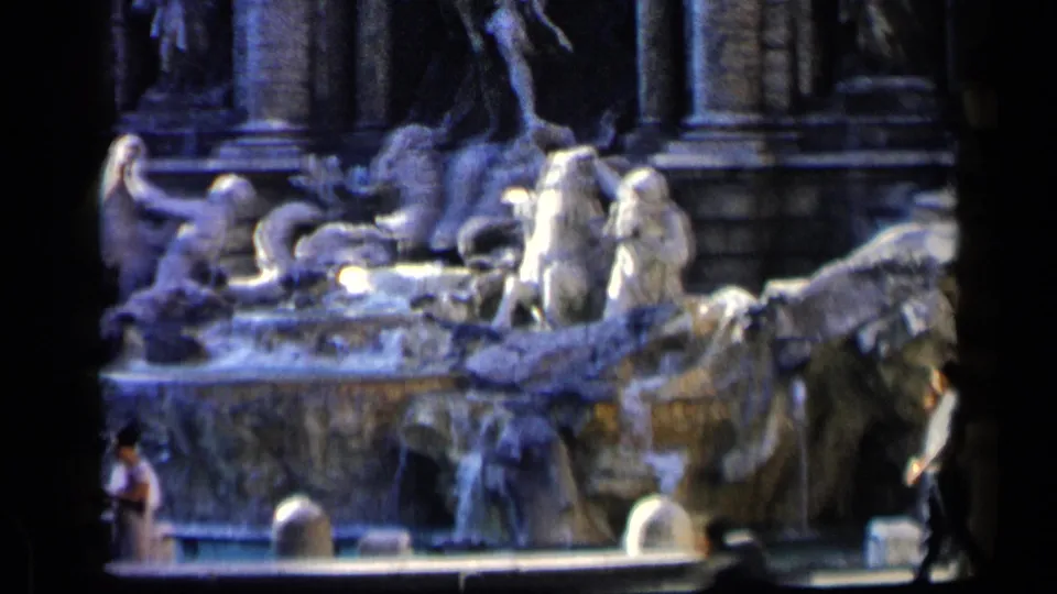 Rome's Trevi Fountain fills the frame, with pale stone figures, water, bollards, and a visitor partly visible at the lower right.