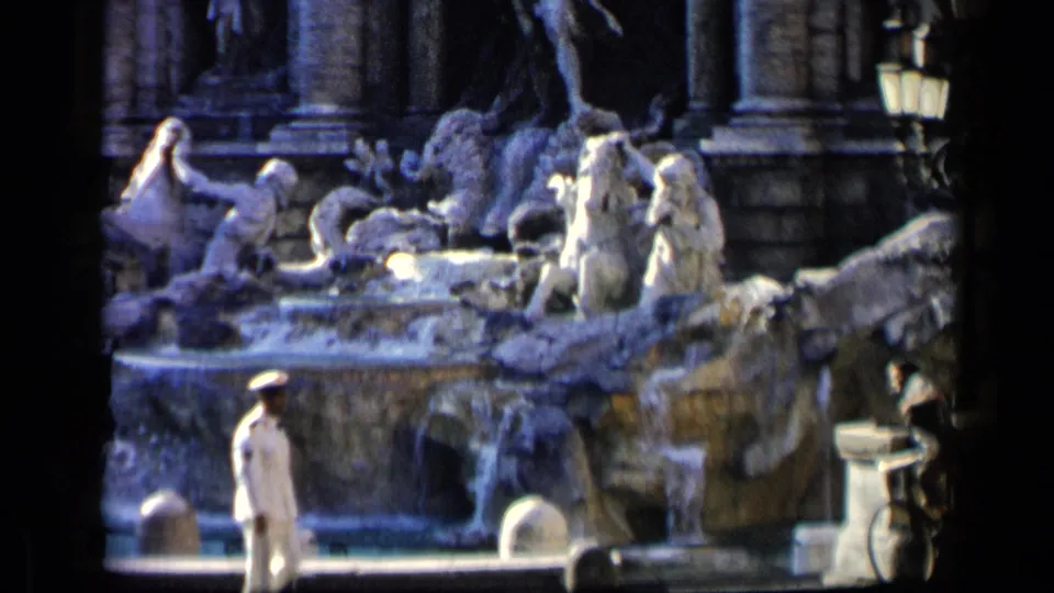 1960 8mm frame showing a white-uniformed man passing before a large sculpted fountain in Italy.